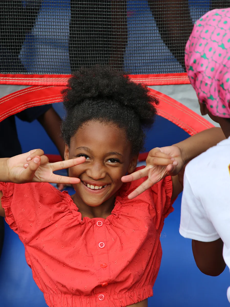 A smiling child in a red shirt making peace signs with both hands, standing near a friend